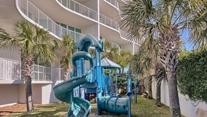 Een speeltuin met een spiraalvormige glijbaan en klimconstructies, omgeven door palmbomen en een gebouw met balkons.