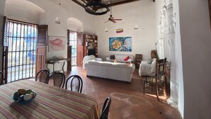 A spacious room with a high ceiling, a ceiling fan, a large window with bars, a dining table with chairs, a white sofa, a bookshelf, and a flag on the wall.