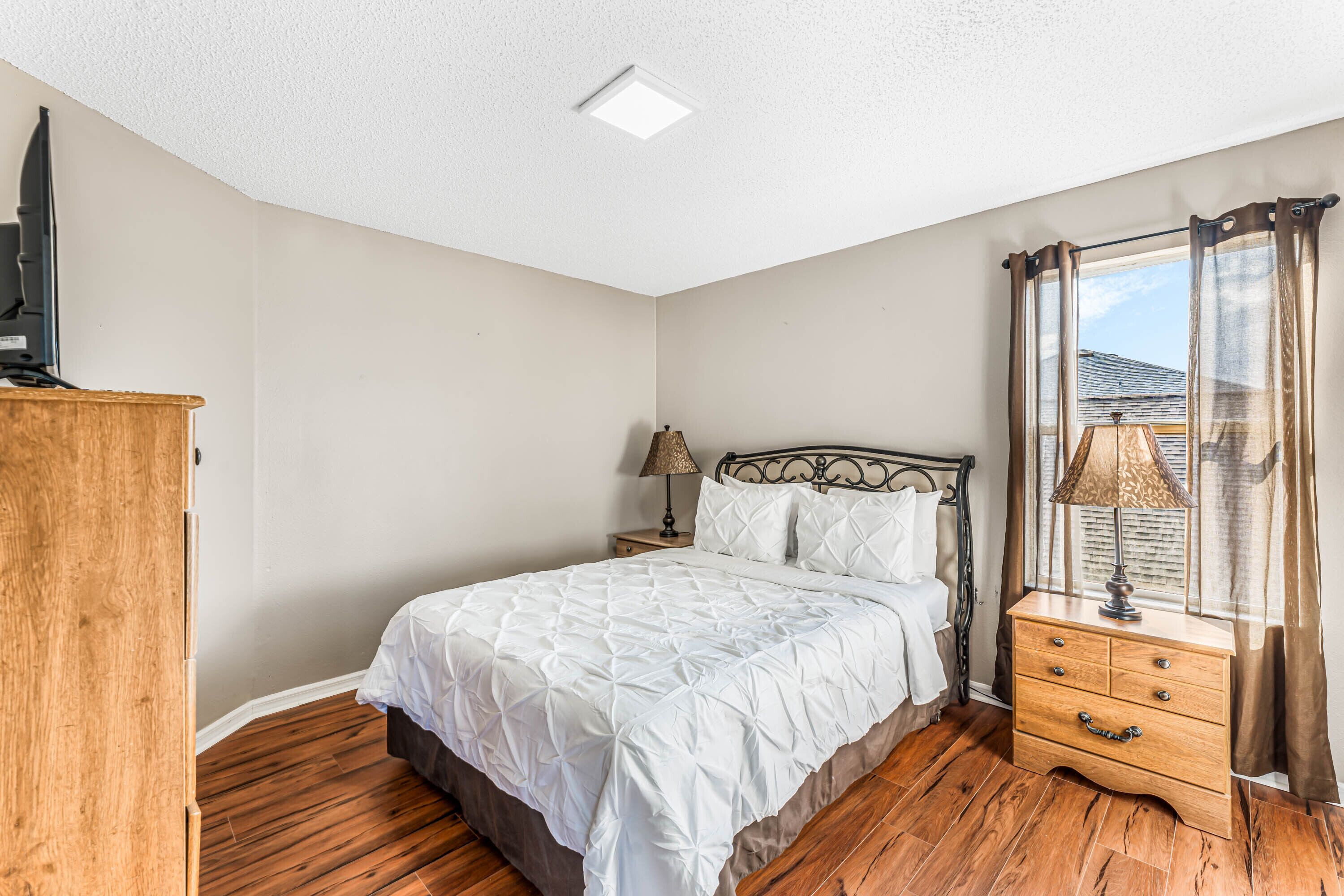 Une chambre à coucher avec un lit, une commode en bois, une table de chevet avec une lampe et une fenêtre avec des rideaux.