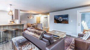 Un salon moderne avec un canapé, une table basse et des tabourets de bar. On y trouve également un coin repas avec des chaises en bois et un lustre. Un tableau est accroché au mur, et un téléviseur est fixé au-dessus de la table basse.