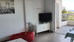 Un salon moderne avec un téléviseur à écran plat, un meuble blanc, un fauteuil poire rouge, une petite table blanche et un balcon avec vue.