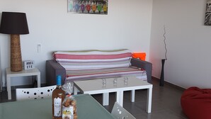 Un salon moderne avec une table à manger pour deux personnes, un canapé, un fauteuil en forme de poire rouge, une lampe et un climatiseur fixé au mur.
