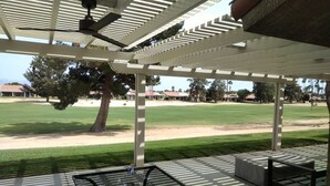 Um espaço coberto ao ar livre com mesa e cadeiras, uma pérgola e vista para um campo verde e edifícios ao fundo.