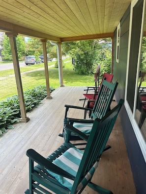 A covered porch with wooden flooring, green rocking chairs, and a view of a green lawn and parked cars.