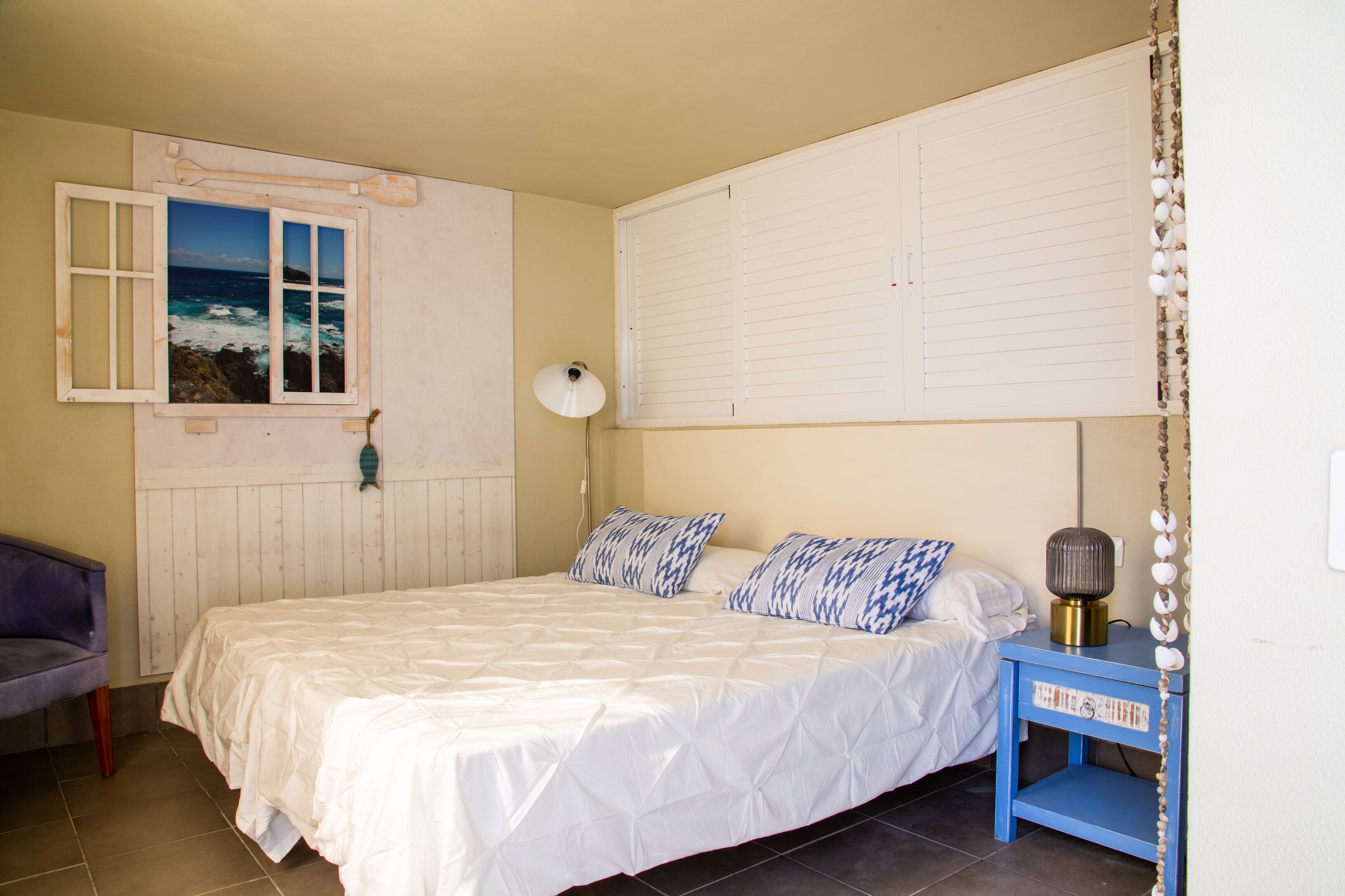 A bedroom with a bed, a nightstand, a lamp, a window with a view of the sea, and a chair.
