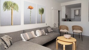 Uma sala de estar moderna com um sofá cinzento, duas cadeiras de vime, uma mesa redonda pequena e três impressões botânicas emolduradas na parede.