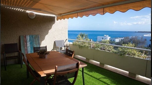 Varanda com mesa e cadeiras de madeira, um vaso com planta e vista para o mar e edifícios.
