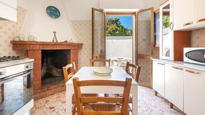 Een keuken met een bakstenen open haard, een eettafel met stoelen, een klok aan de muur en keukenkastjes.