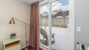 Een kamer met een schuifdeur die uitkomt op een besneeuwde buitenruimte, een vloerlamp, een kleine tafel met een plant en een bankje buiten.