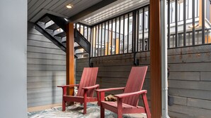 Een overdekt terras met twee rode Adirondack-stoelen, een geboeide vloerkleden en een uitzicht op een trap en gebouwen op de achtergrond.