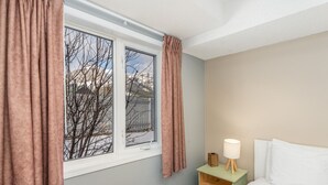 Een slaapkamer met een bed, een nachtkastje met een lamp, een raam met gordijnen en uitzicht op kale bomen.