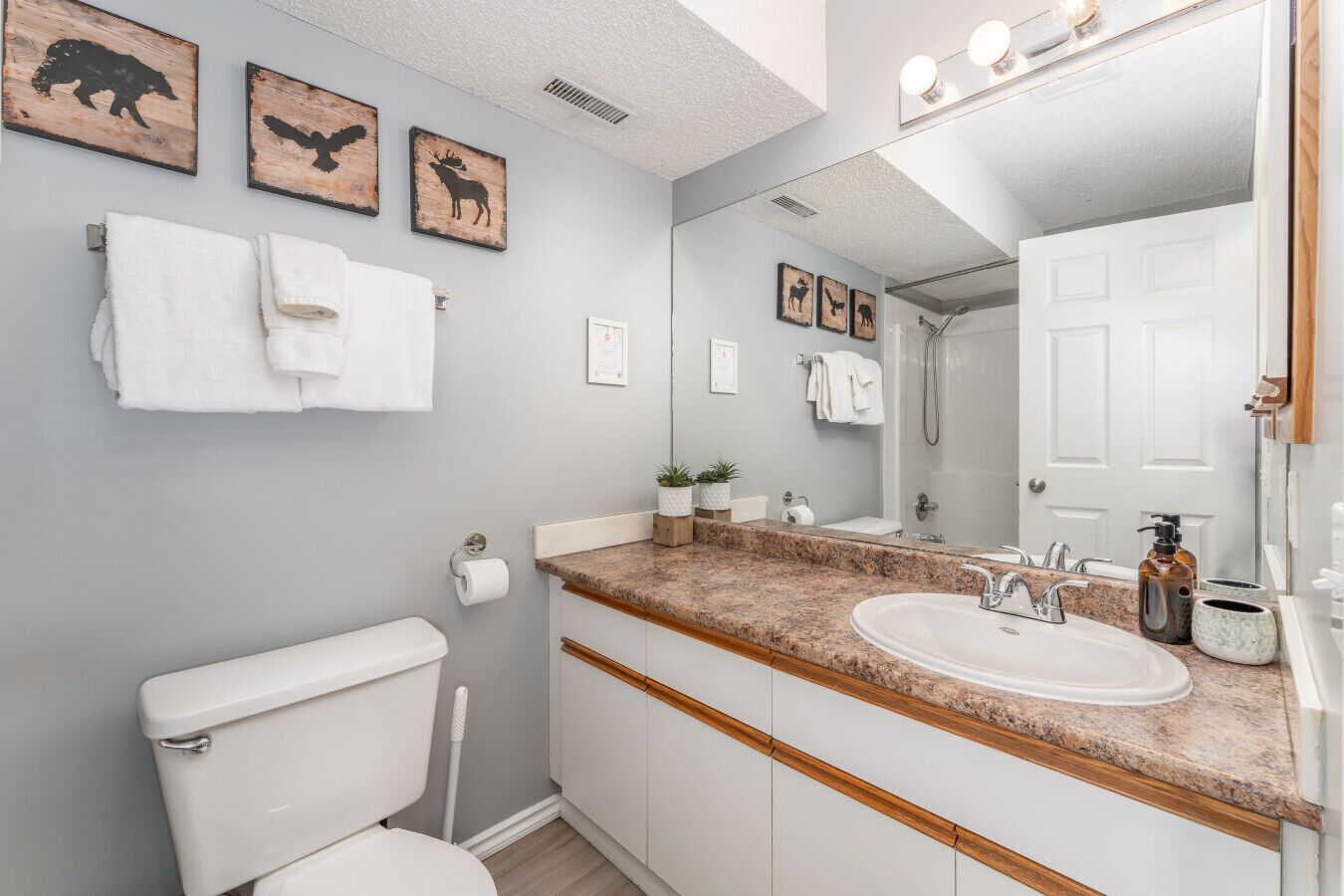 Une salle de bain moderne avec des toilettes blanches, un lavabo doté d’un plan de travail en granit et un grand miroir. Des tableaux encadrés ornent le mur, et un porte-serviettes est muni de serviettes blanches.