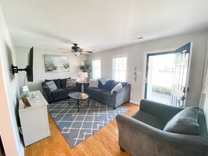 A living room with two sofas, a coffee table, a TV mounted on the wall, a ceiling fan, and a door leading outside.