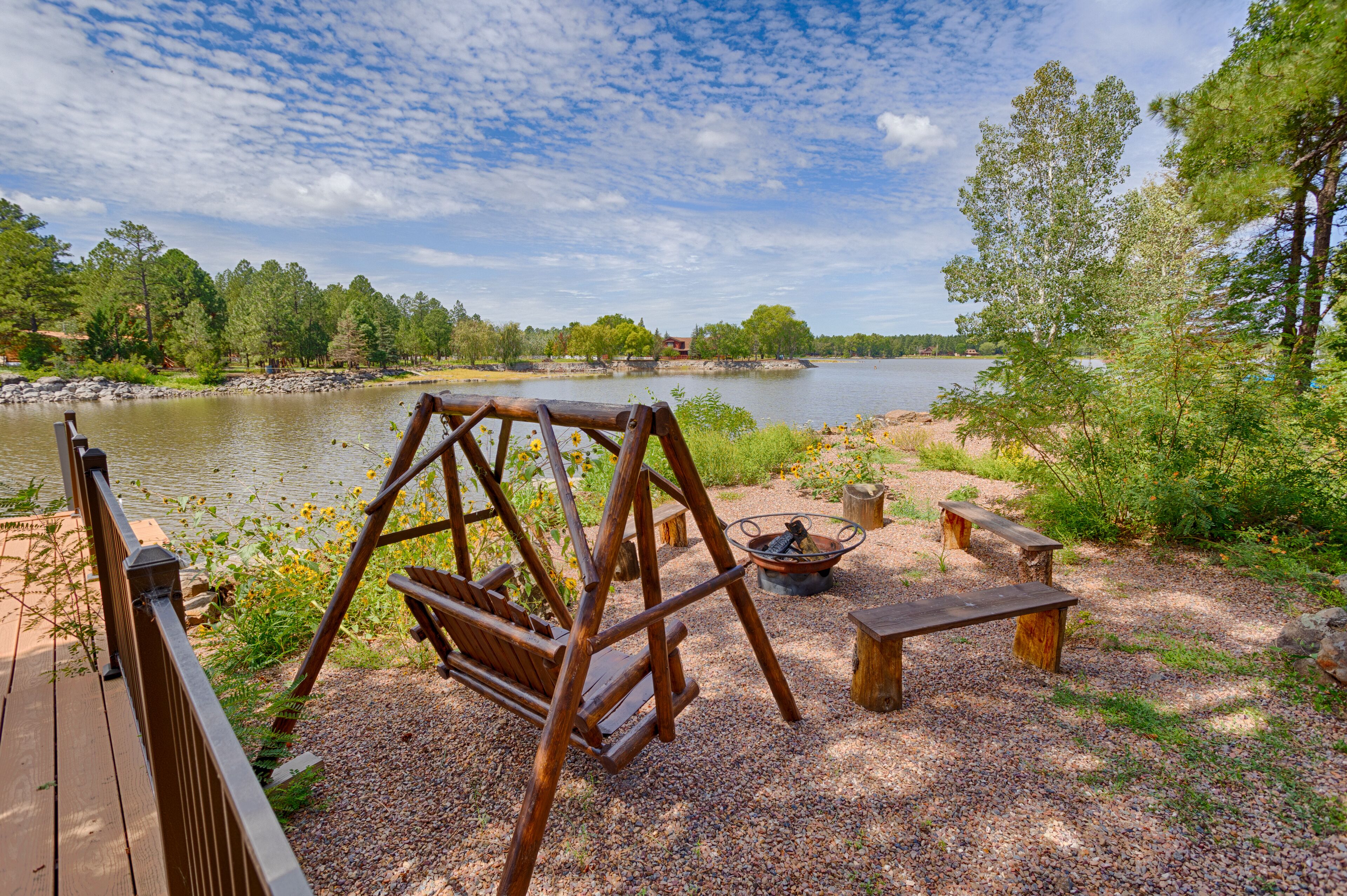 A wooden swing set, a fire pit, and a bench by a lake with a wooden deck.