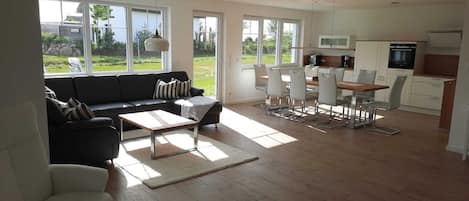 A spacious living room with a sofa, coffee table, and dining area with chairs. Large windows provide natural light and a view of the outdoors.