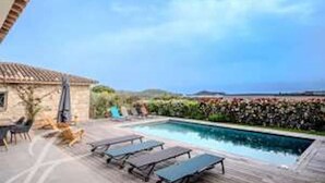 Une piscine avec des chaises longues, un bâtiment en pierre et une vue sur la mer.