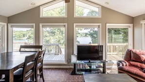 A spacious room with a high ceiling, large windows, a dining area with a table and chairs, a glass coffee table, a leather sofa, and a flat-screen TV. - Lakeside Cottage w/ Spacious Multi-level Deck and Outdoor Living Space (Monticello)
