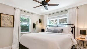 Un dormitorio con cama, ventilador de techo, ventana con cortinas, mesita de noche con lámpara y un cuadro en la pared.