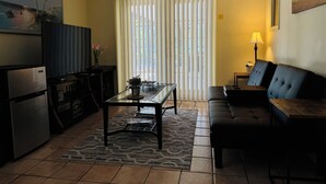 A living room with a black sofa, a glass coffee table, a kitchen area with a refrigerator, and a large window with blinds.