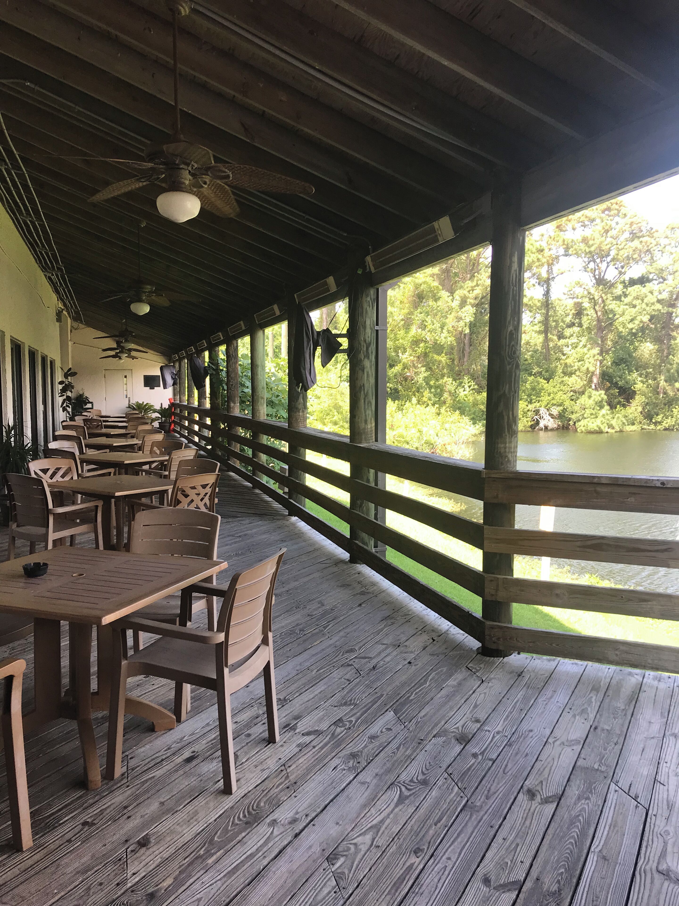 A covered outdoor seating area with wooden tables and chairs, overlooking a body of water and surrounded by greenery.