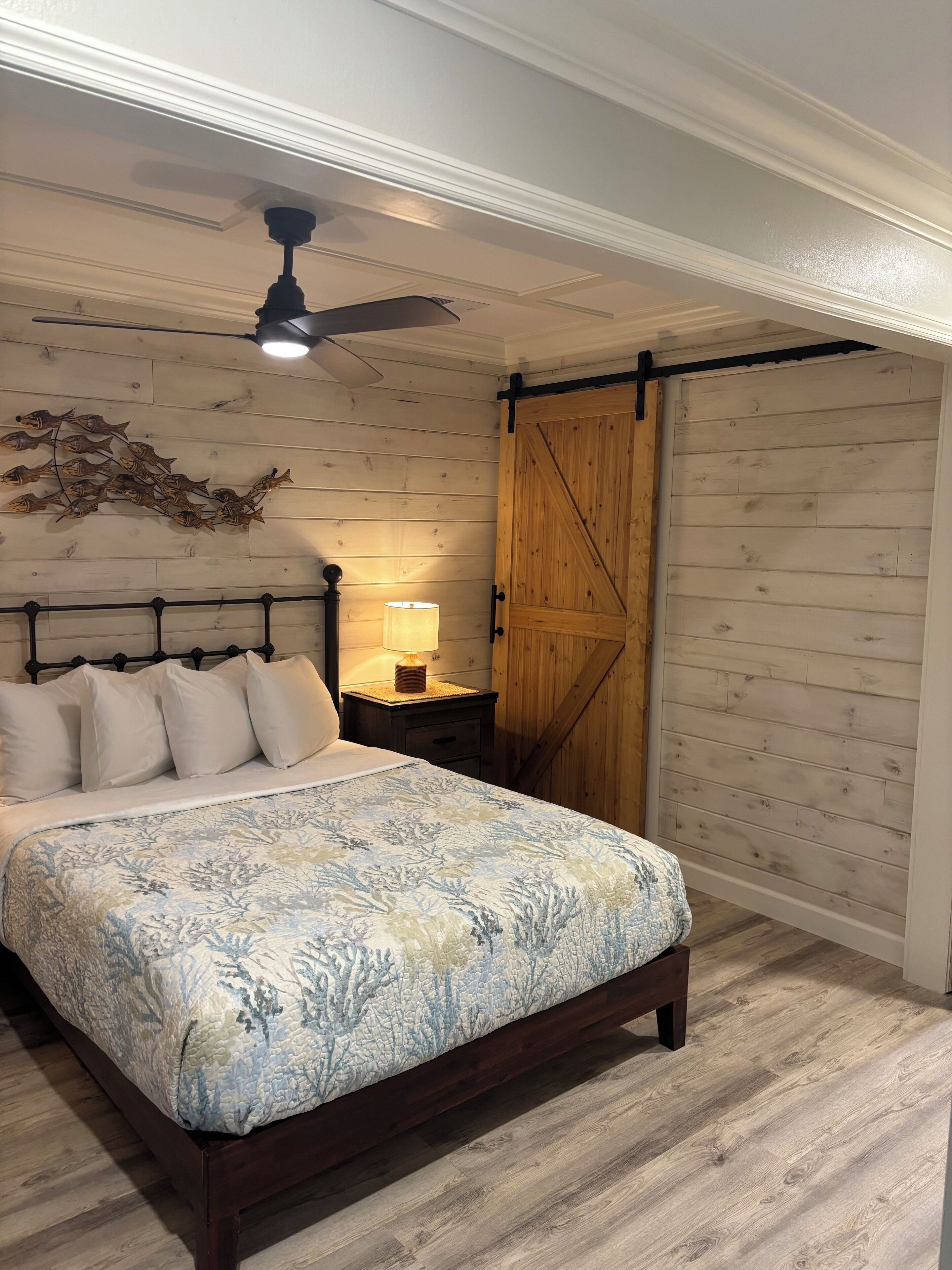 A bedroom with a bed, a wooden headboard, a ceiling fan, a wall-mounted lamp, and a wooden door.