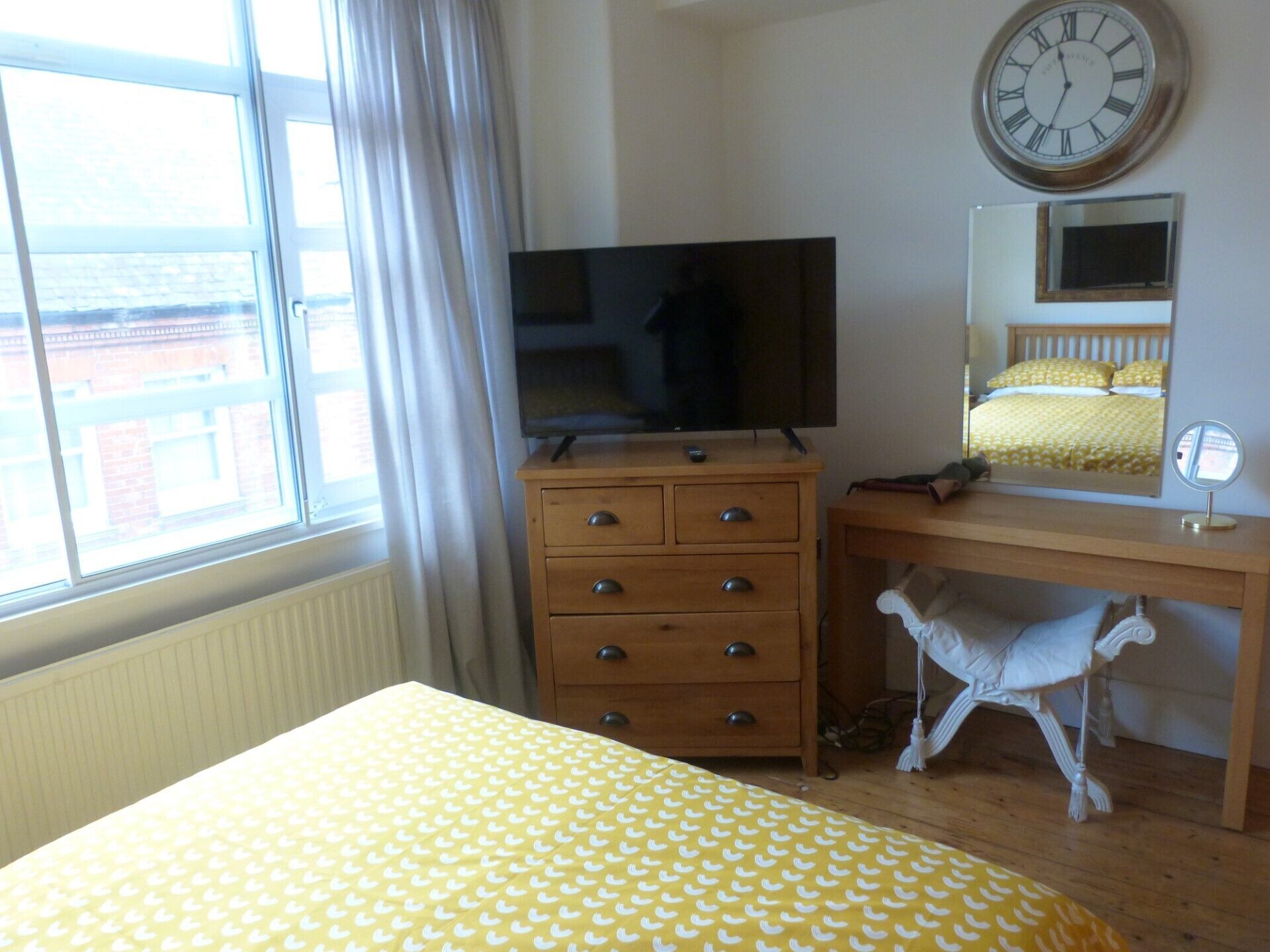 Ein Schlafzimmer mit einem Bett, einer Holzkommode, einem Fernseher, einem Spiegel und einem Fenster mit Vorhängen.