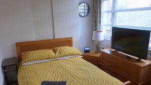 Una cama bien hecha con una colcha amarilla estampada, una lámpara de noche, un televisor sobre un soporte de madera y una mesita de noche.