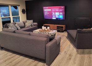 Un salon moderne avec un canapé d’angle gris, une table basse agrémentée de bougies et un téléviseur à écran plat fixé au mur, affichant une interface de la marque TCL.