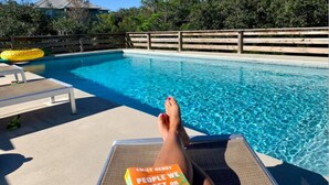 A poolside area with a lounge chair, a book titled "People We Meet on Vacation," and a wooden fence. - WALK TO PRIVATE ACCESS  "PET FRIENDLY BEACH"  ENJOY 
PRIVATE POOL & HOT TUB (Gulf Shores)