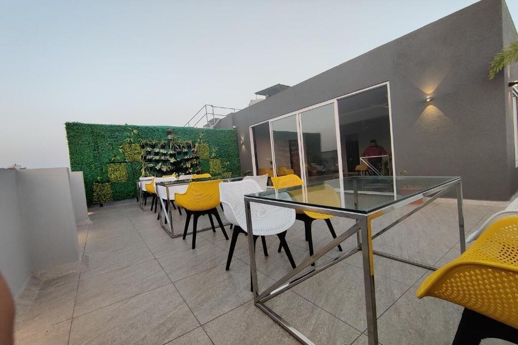 An outdoor patio with a glass table and chairs, a green wall with plants, and a modern building in the background.