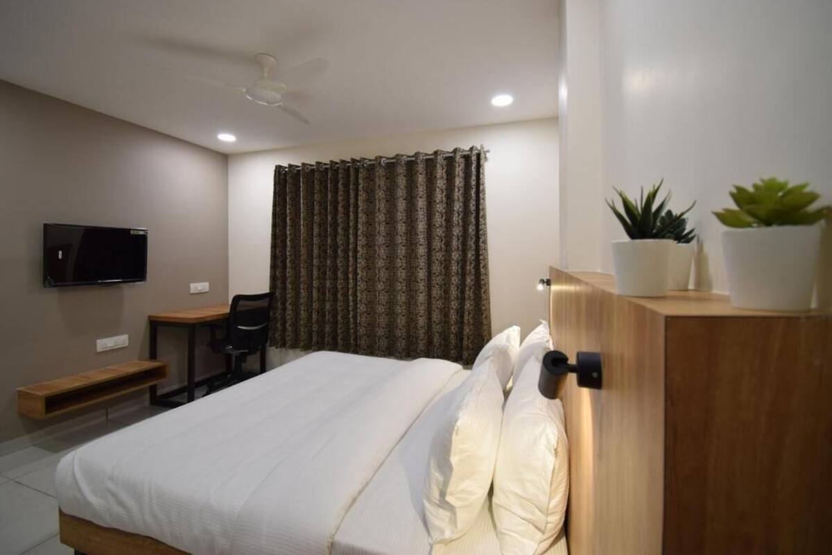 A hotel room with a bed, a desk, a chair, a television, a ceiling fan, and two potted plants.