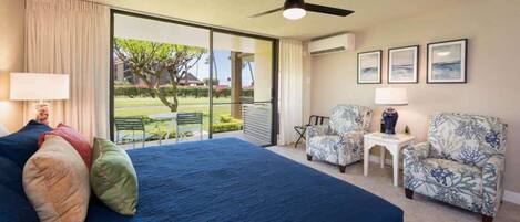 Habitación de hotel con una cama grande, un ventilador de techo, vistas al exterior, una mesita con lámpara y dos sillones estampados.