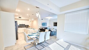 Una cocina moderna con armarios blancos, un comedor con mesa dispuesta para cuatro personas y una sala de estar con sofá y televisor.