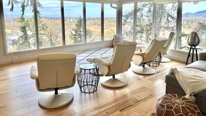Ruime kamer met grote ramen, met uitzicht op een bos en gebouwen in de verte. Drie stoelen en een kleine ronde tafel.