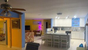 A spacious living area with a dining table, chairs, and a ceiling fan. A kitchen with white cabinets and a tiled backsplash is visible in the background.