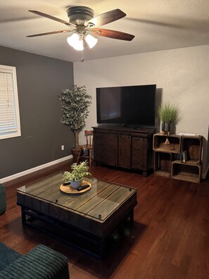 Una sala de estar con ventilador de techo, televisor de pantalla plana, mesa de centro de madera, una planta y una estantería.