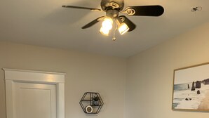 Une chambre à coucher comprenant un lit, un ventilateur de plafond, une horloge murale, un tableau encadré et une petite table sur laquelle se trouvent une plante et une lampe.