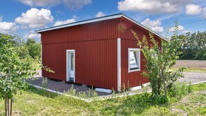Punainen, yhden kerroksen korkuinen rakennus, jossa on kaksi ovea ja yksi ikkuna. Rakennuksen ympärillä on vehreää kasvillisuutta ja taivaalla ei ole pilviä.