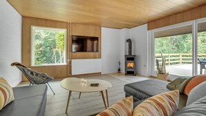 Un salon moderne avec un plafond en bois, une cheminée, un téléviseur fixé au mur et de grandes fenêtres offrant une vue sur l’extérieur.