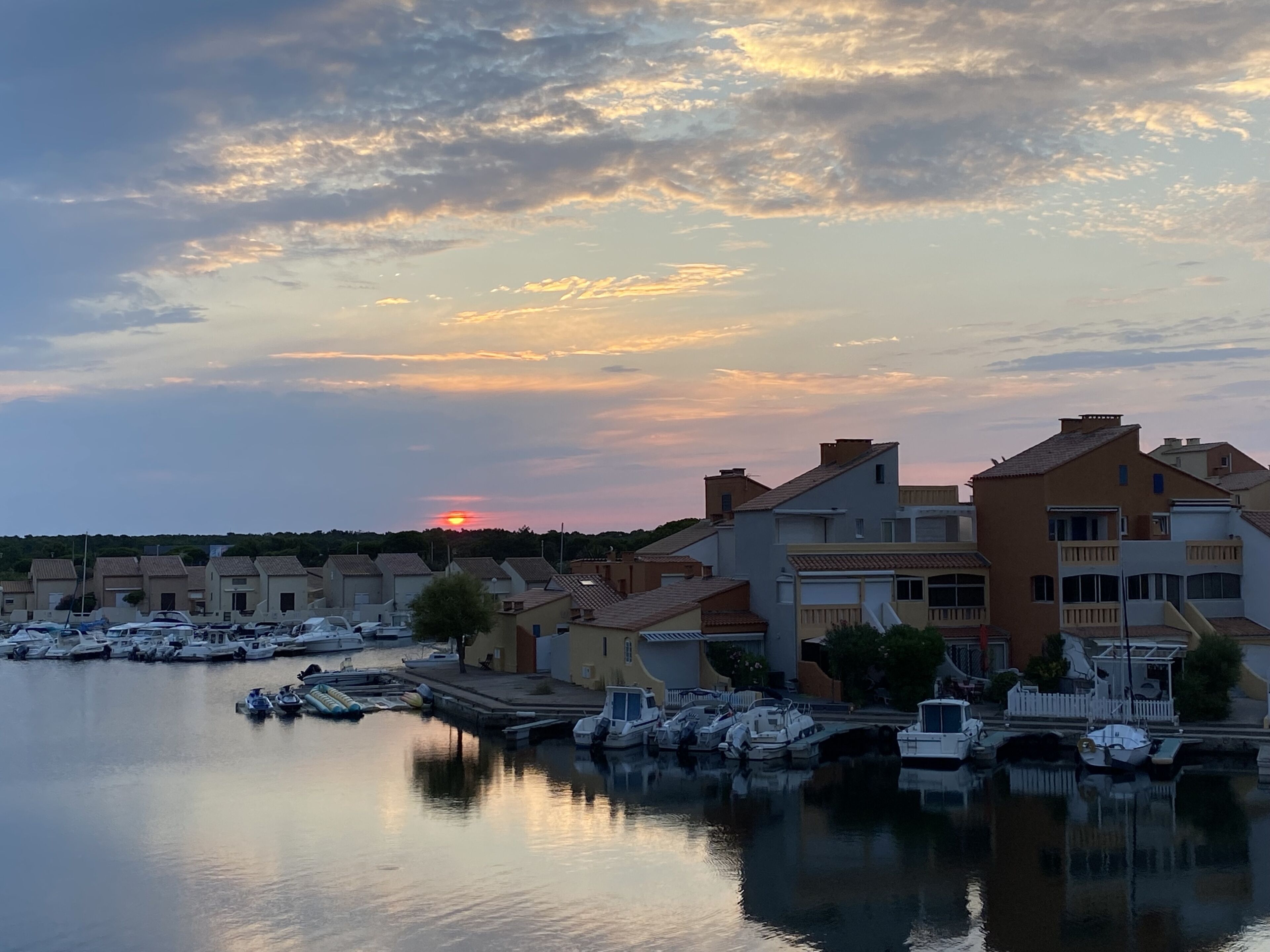 Ein Yachthafen mit vertäuten Booten, ein Sonnenuntergang im Hintergrund und Wohngebäude mit Balkonen.