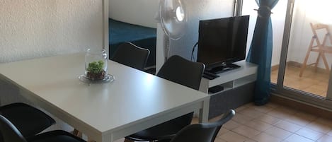 A dining area with a white table, black chairs, a small plant, a fan, and a TV.