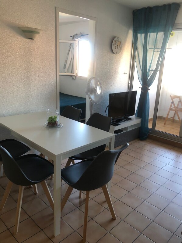 A dining area with a white table, black chairs, a small plant, a fan, and a TV.