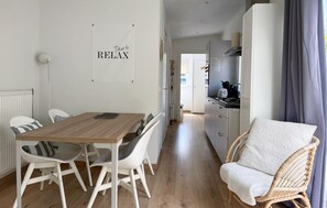 Eine moderne Küche mit weißen Schränken, ein Essbereich mit einem Holztisch und weißen Stühlen sowie ein Korbstuhl mit weißem Sitzkissen.