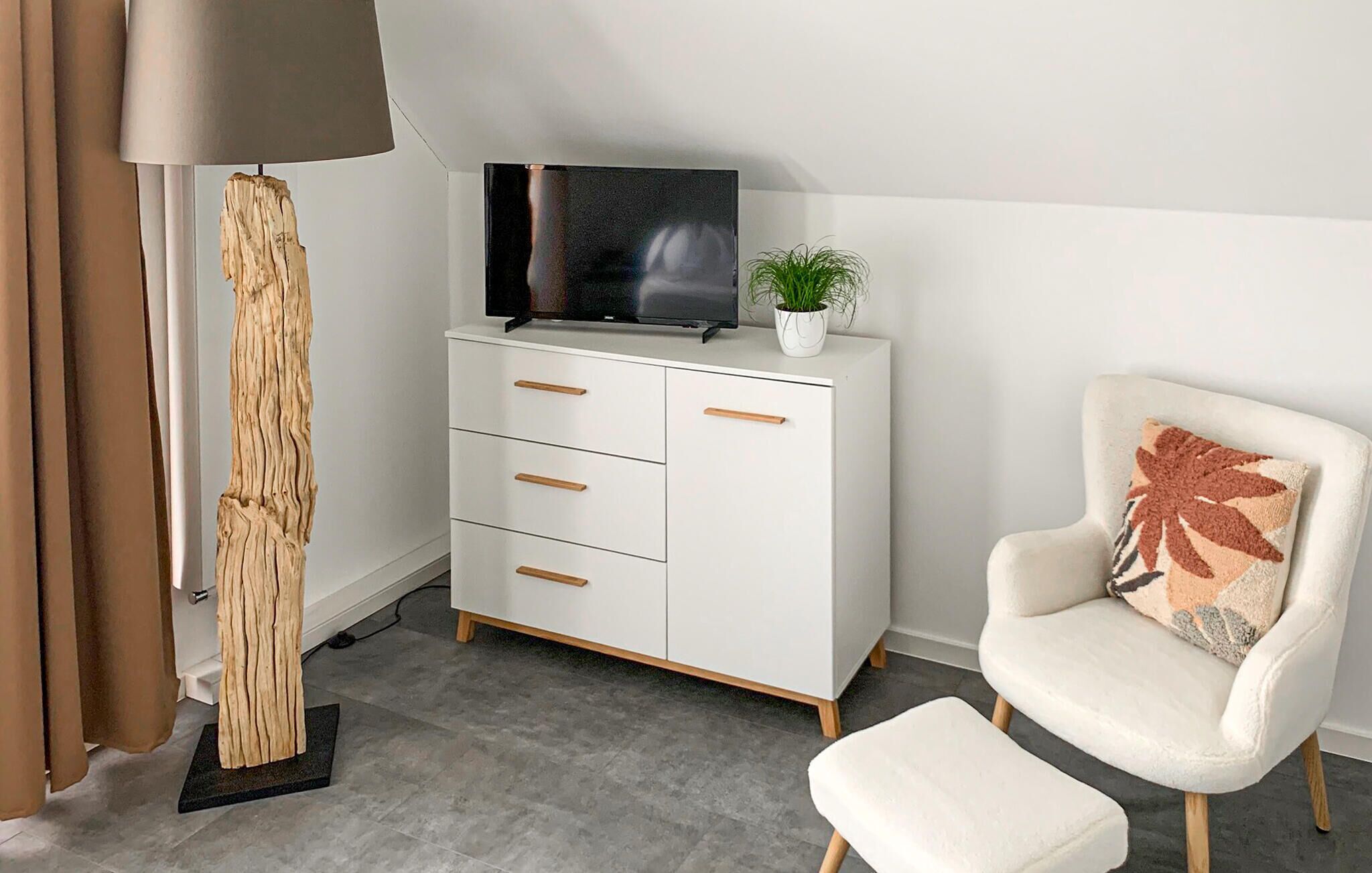 A modern living room with a white cabinet, a flat-screen TV, a potted plant, a white armchair with a patterned cushion, and a tall wooden floor lamp.