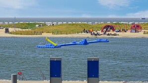 Una zona costera con un muelle, estructuras azules flotantes y una playa con gente y una estructura roja al fondo.