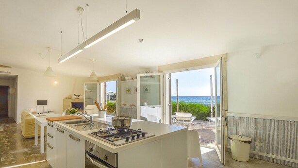 A modern kitchen with a central island, a dining area with a view of the ocean, and a large sliding glass door leading to an outdoor patio. - 3 bedroom beach front home in Siracusa (Siracusa)