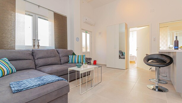 Una sala moderna con un sofá gris, una mesita de centro y una silla negra. Se observa también una cocina con una encimera y un espejo.