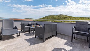 Terraza en la azotea con mobiliario de mimbre y una mesa, con vistas a una colina verde.