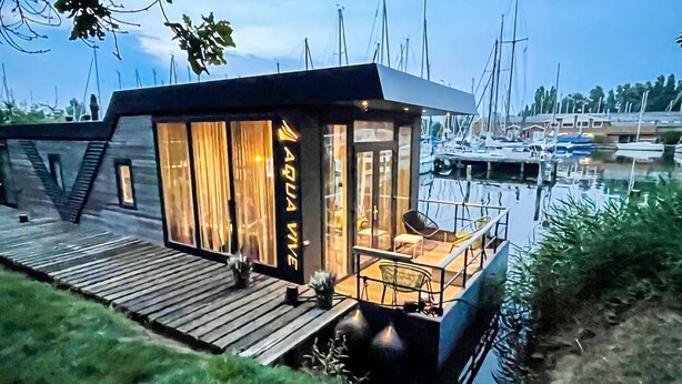 Ein modernes Hausboot mit Glasfassade, Holldeck und Blick auf den Hafen.