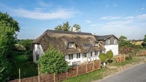 A traditional house with a thatched roof and white walls, surrounded by lush greenery and a wooden fence. - Nice home in Søllested with sauna (Søllested)